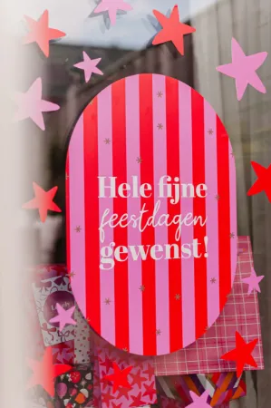 Sfeerbeeld raamsticker 40x60cm ovale vorm met roze-rode kleuren en tekst "Hele fijne feestdagen gewenst" op een raam in de etalage